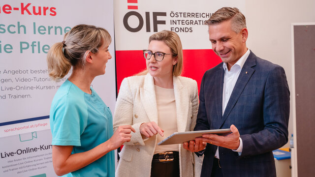 Drei Personen unterhalten sich und betrachten gemeinsam ein Tablet, während ein Banner im Hintergrund sichtbar ist.