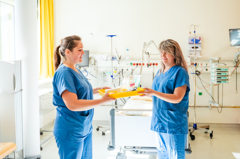 Zwei Krankenschwestern übergeben sich einen gelben Tablett mit medizinischen Utensilien in einem Krankenhauszimmer.