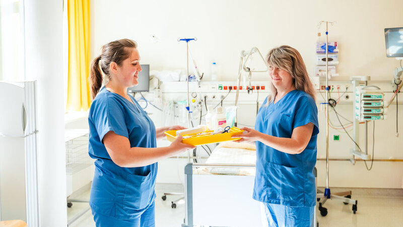 Zwei Krankenschwestern übergeben sich einen gelben Tablett mit medizinischen Utensilien in einem Krankenhauszimmer.