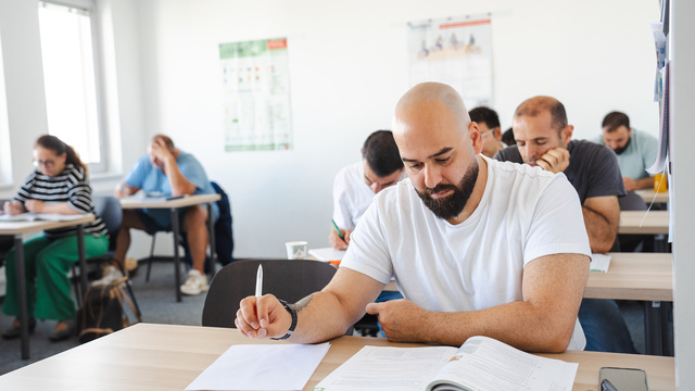 In einem Klassenzimmer sitzen mehrere Erwachsene an Tischen und arbeiten konzentriert. Ein Mann mit Glatze und Bart schreibt in ein Heft und blickt auf ein Lehrbuch. Andere Teilnehmer sind im Hintergrund ebenfalls mit Aufgaben beschäftigt.