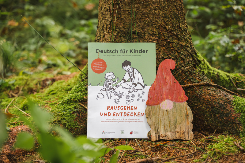 Ein Plakat mit dem Titel "Deutsch für Kinder", das die Aktivitäten im Freien fördert, neben einer Holzfigur eines Wichtels.