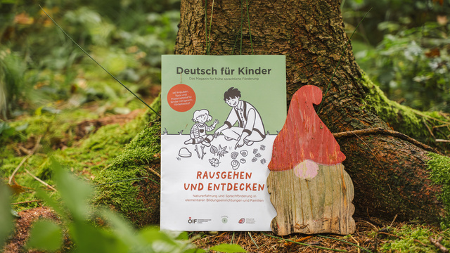 Ein Plakat mit dem Titel "Deutsch für Kinder", das die Aktivitäten im Freien fördert, neben einer Holzfigur eines Wichtels.