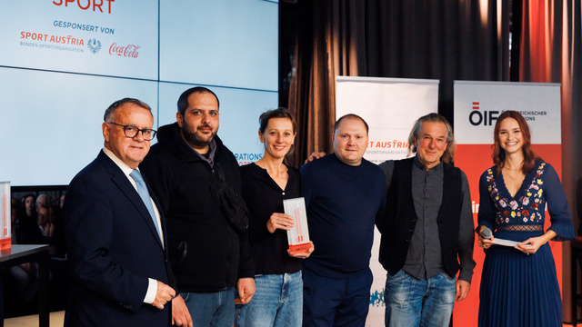 Eine Gruppe von fünf Personen steht bei einer Sportveranstaltung vor einer großen Bildschirmwand.