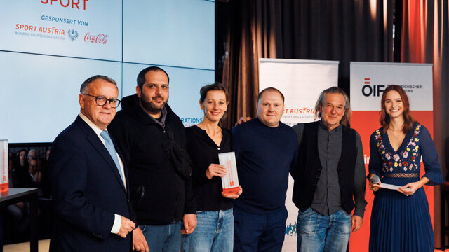 Eine Gruppe von fünf Personen steht bei einer Sportveranstaltung vor einer großen Bildschirmwand.