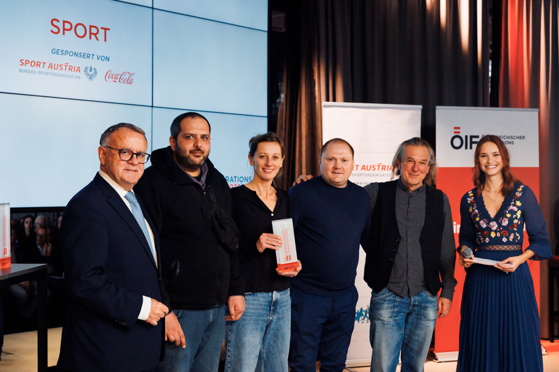 Eine Gruppe von fünf Personen steht bei einer Sportveranstaltung vor einer großen Bildschirmwand.