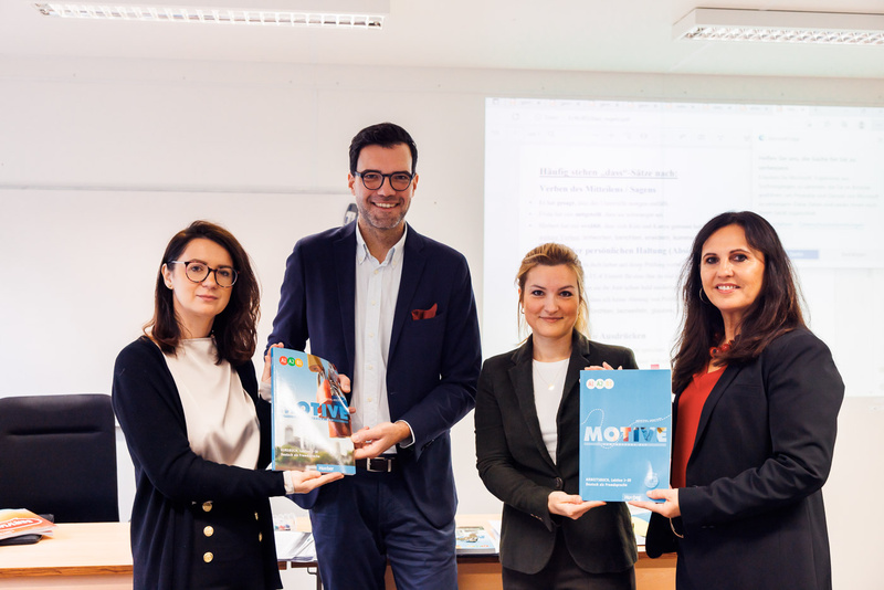 Vier Personen halten Bücher in der Hand, stehen vor einem Tisch in einem hellen Raum.