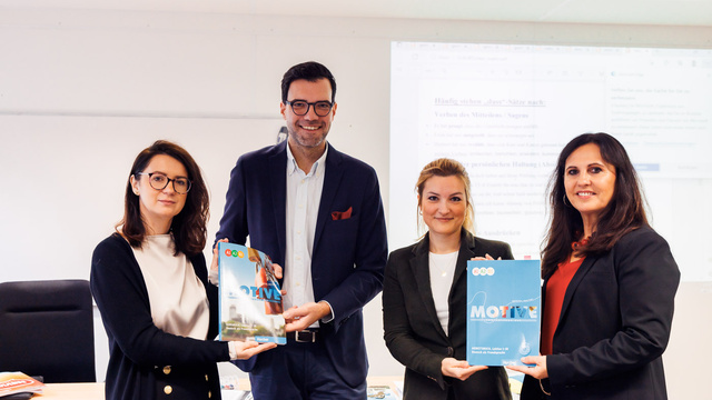 Vier Personen halten Bücher in der Hand, stehen vor einem Tisch in einem hellen Raum.