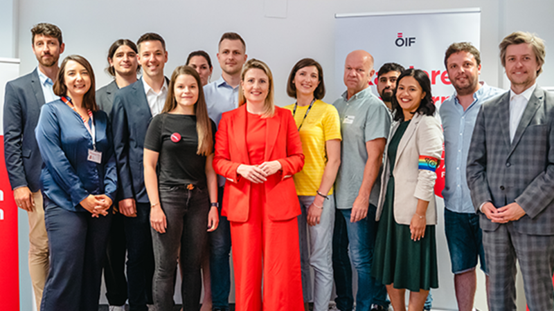 Eine Gruppe von zwölf Personen steht zusammen, lächelnd, in einem Raum mit einem OIF-Logo im Hintergrund.