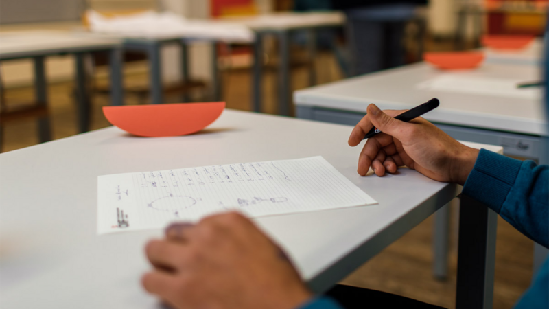 Eine Hand hält einen Stift über einem Arbeitsblatt auf einem Tisch in einem Klassenzimmer.