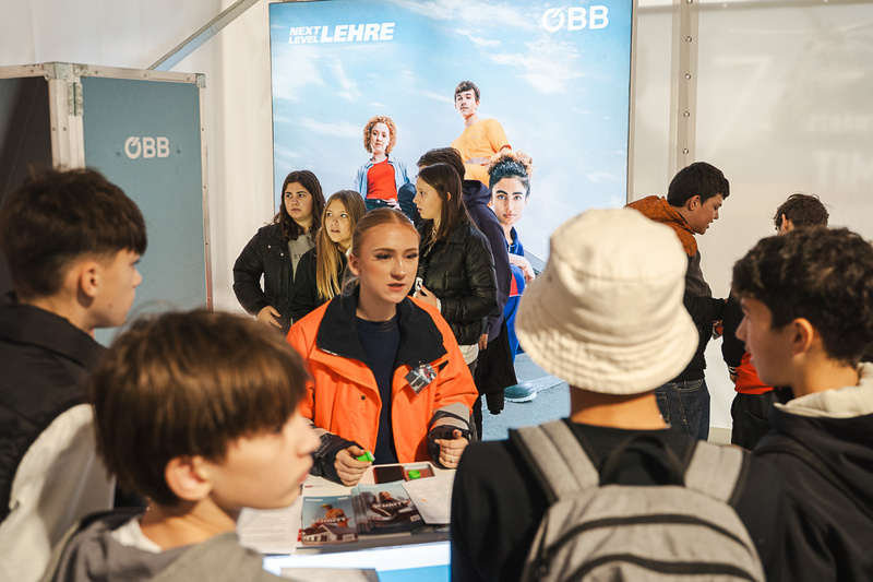 Junge Erwachsene stehen an einem Stand, während eine Mitarbeiterin Informationen teilt.