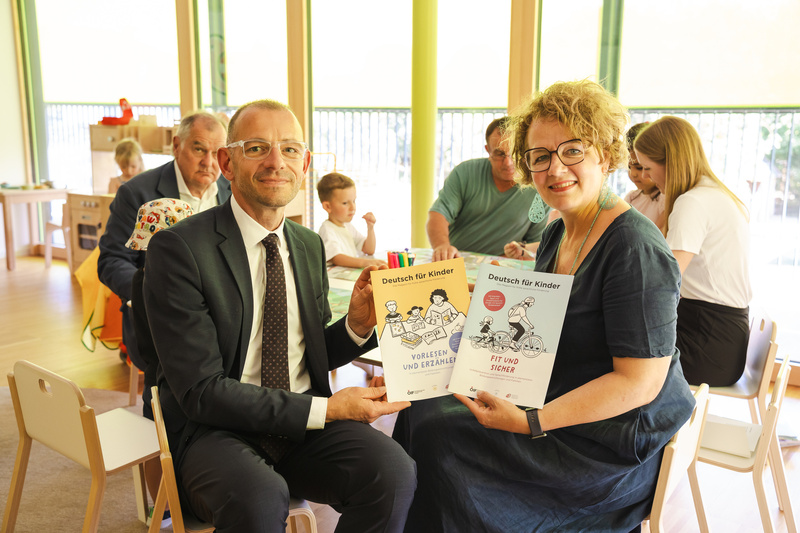 Ein Mann und eine Frau halten Bücher mit dem Titel "Deutsch für Kinder" in einem hellen Raum.