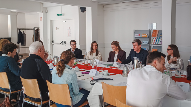 Eine Gruppe von Personen sitzt an einem langen Tisch in einem Besprechungsraum. Einige Teilnehmer diskutieren, während andere zuhören. Auf dem Tisch stehen Kaffeekannen und Notizmaterialien. Der Raum ist hell und modern eingerichtet.