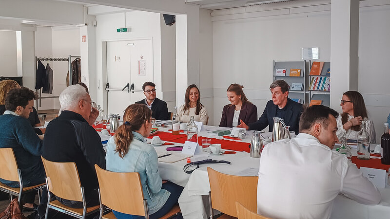 Eine Gruppe von Personen sitzt an einem langen Tisch in einem Besprechungsraum. Einige Teilnehmer diskutieren, während andere zuhören. Auf dem Tisch stehen Kaffeekannen und Notizmaterialien. Der Raum ist hell und modern eingerichtet.