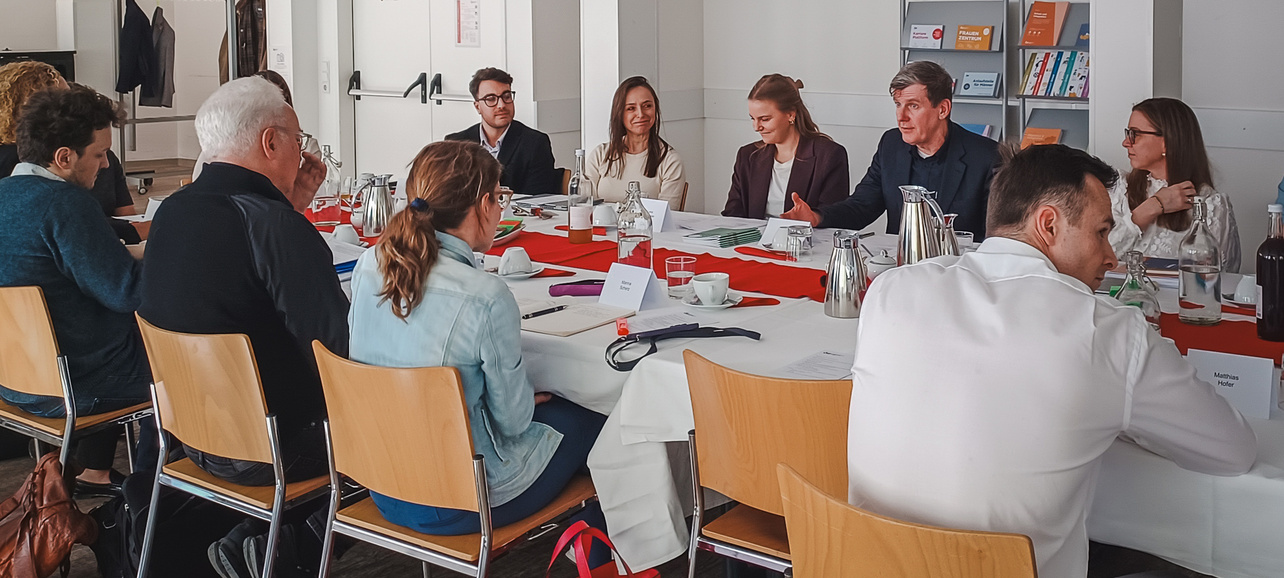 Eine Gruppe von Personen sitzt an einem langen Tisch in einem Besprechungsraum. Einige Teilnehmer diskutieren, während andere zuhören. Auf dem Tisch stehen Kaffeekannen und Notizmaterialien. Der Raum ist hell und modern eingerichtet.