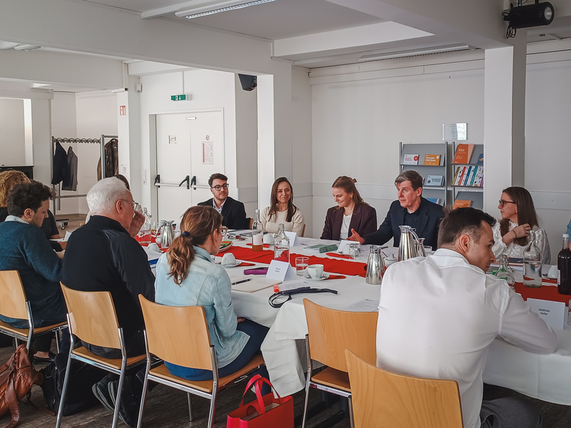 Eine Gruppe von Personen sitzt an einem langen Tisch in einem Besprechungsraum. Einige Teilnehmer diskutieren, während andere zuhören. Auf dem Tisch stehen Kaffeekannen und Notizmaterialien. Der Raum ist hell und modern eingerichtet.
