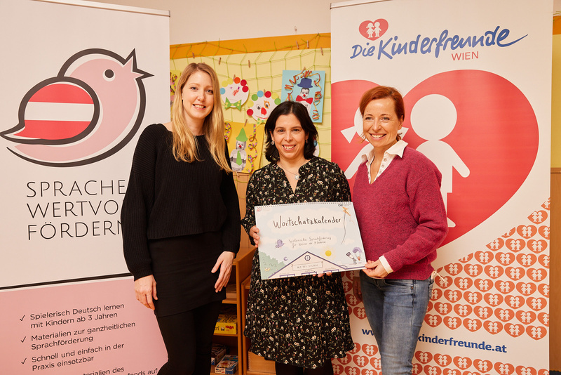 Drei Frauen halten einen Kalender für sprachliche Förderung in der Hand, im Hintergrund sind Banner zu sehen.