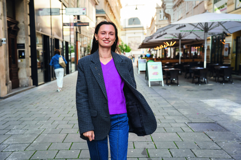 Junge Frau in buntem Pullover und Blazer lächelt, während sie entspannt eine Straße entlanggeht.