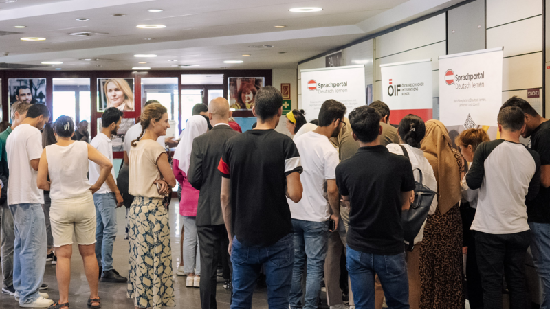 Eine Gruppe von Menschen steht an Informationsständen für Deutschkurse in einem öffentlichen Raum.