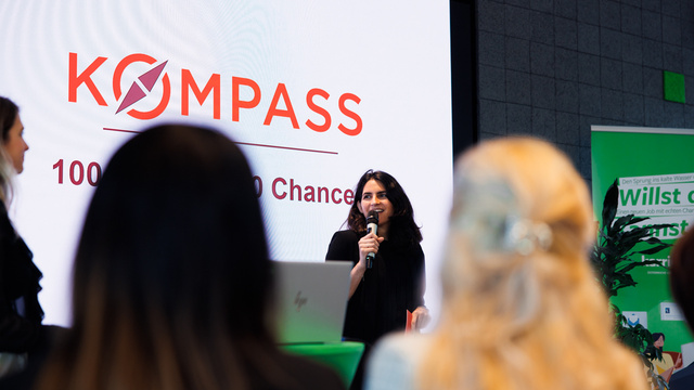 Eine Frau spricht auf einer Veranstaltung mit dem Titel "KOMPASS – 100 Chancen für 100 Chancen".