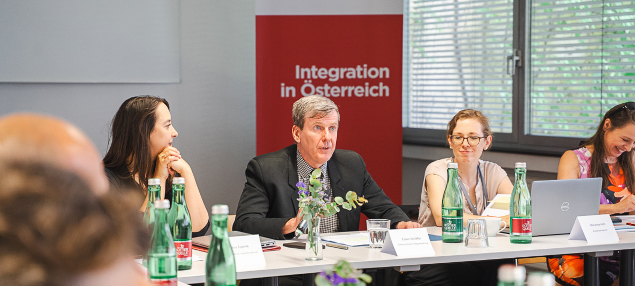 Eine Gruppe von Menschen sitzt an einem Tisch in einem Besprechungsraum. Ein Mann in einem Anzug spricht, während andere Teilnehmer, darunter Frauen, aufmerksam zuhören. Im Hintergrund steht ein Banner mit der Aufschrift "Integration in Österreich". Wasserflaschen und Notizen liegen auf dem Tisch.