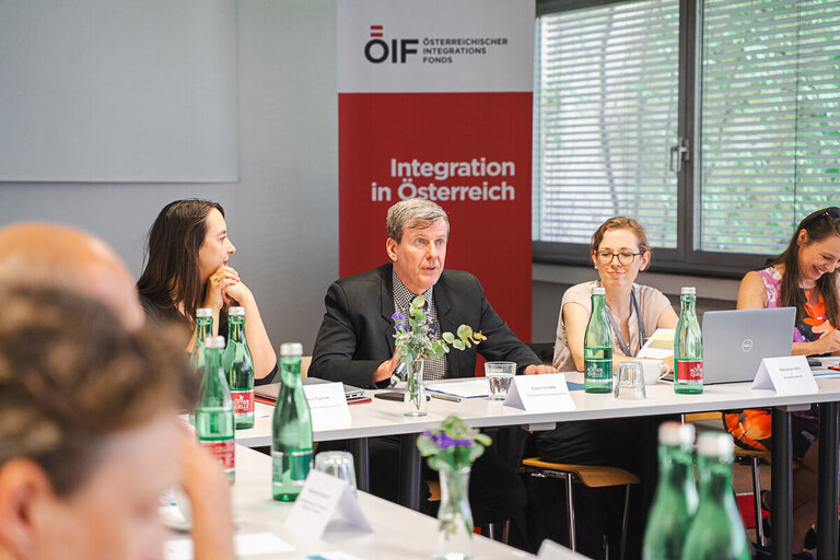 Eine Gruppe von Menschen sitzt an einem Tisch in einem Besprechungsraum. Ein Mann in einem Anzug spricht, während andere Teilnehmer, darunter Frauen, aufmerksam zuhören. Im Hintergrund steht ein Banner mit der Aufschrift 