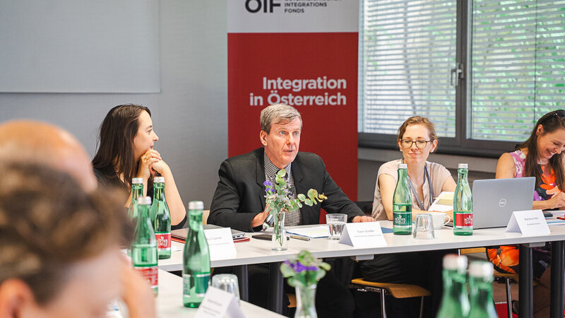 Eine Gruppe von Menschen sitzt an einem Tisch in einem Besprechungsraum. Ein Mann in einem Anzug spricht, während andere Teilnehmer, darunter Frauen, aufmerksam zuhören. Im Hintergrund steht ein Banner mit der Aufschrift "Integration in Österreich". Wasserflaschen und Notizen liegen auf dem Tisch.