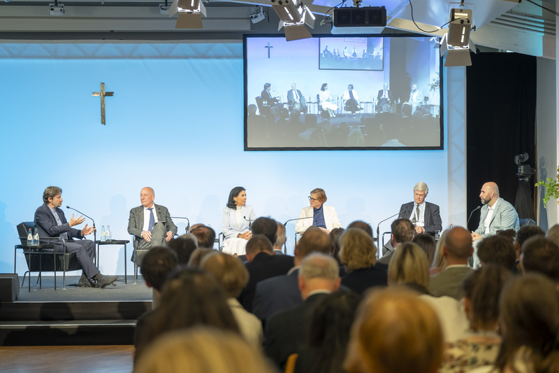 Podiumsdiskussion mit sechs Teilnehmenden, die an einem Tisch sitzen. Im Hintergrund ist eine Leinwand mit einer Präsentation zu sehen. Das Publikum ist in der unteren Bildhälfte sichtbar. Ein Kreuz hängt an der Wand.