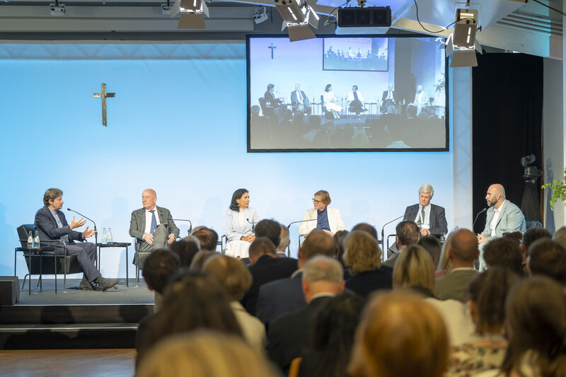 Podiumsdiskussion mit sechs Teilnehmenden, die an einem Tisch sitzen. Im Hintergrund ist eine Leinwand mit einer Präsentation zu sehen. Das Publikum ist in der unteren Bildhälfte sichtbar. Ein Kreuz hängt an der Wand.