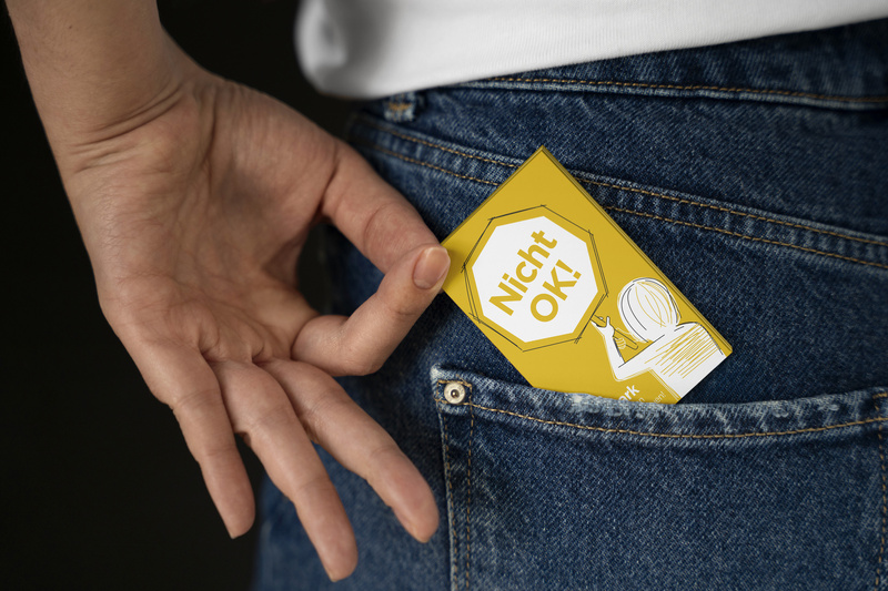 Eine Hand greift nach einer gelben Karte mit dem Aufdruck "Nicht OK!" in einer Jeans-Tasche.