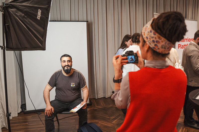 Ein Mann sitzt auf einem Stuhl und wird von einer Person mit einer Kamera fotografiert.