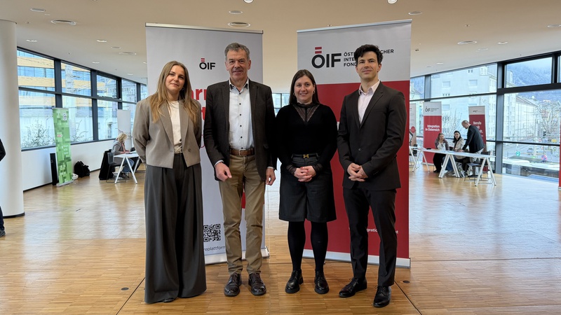 Vier Personen stehen in einem hellen Raum mit großen Fenstern. Sie lächeln und sind formell gekleidet. Im Hintergrund sind zwei Banner sichtbar, die das Logo einer Organisation zeigen. Der Raum wirkt modern und einladend.