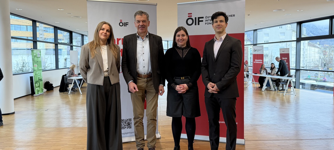 Vier Personen stehen in einem hellen Raum mit großen Fenstern. Sie lächeln und sind formell gekleidet. Im Hintergrund sind zwei Banner sichtbar, die das Logo einer Organisation zeigen. Der Raum wirkt modern und einladend.