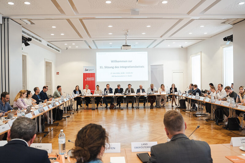 Ein großer Konferenzraum mit einem runden Tisch, an dem viele Teilnehmer sitzen. Auf einem Bildschirm im Hintergrund steht "Willkommen zur 31. Sitzung des Integrationsbeirats". Die Atmosphäre wirkt formal und geschäftlich, mit verschiedenen Personen, die miteinander sprechen und diskutieren.