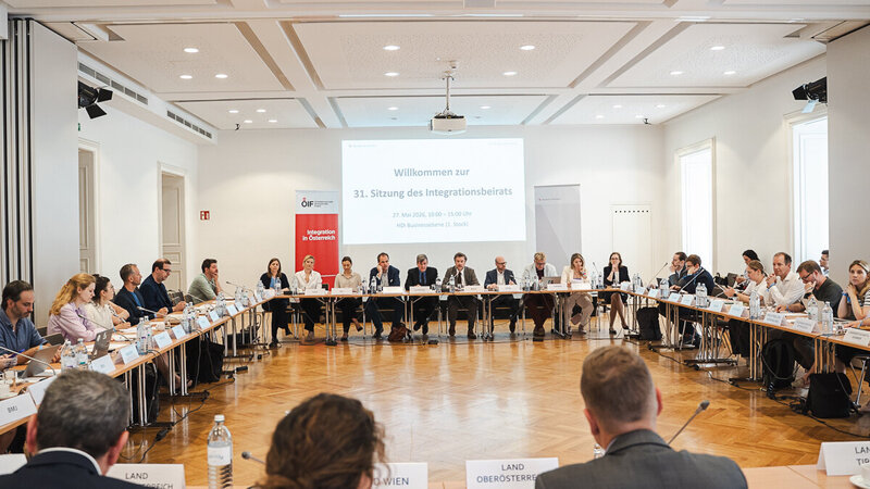 Ein großer Konferenzraum mit einem runden Tisch, an dem viele Teilnehmer sitzen. Auf einem Bildschirm im Hintergrund steht "Willkommen zur 31. Sitzung des Integrationsbeirats". Die Atmosphäre wirkt formal und geschäftlich, mit verschiedenen Personen, die miteinander sprechen und diskutieren.