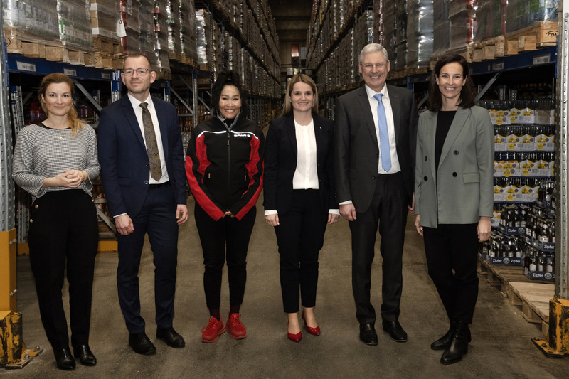 Fünf Personen stehen in einem Lagerhaus zwischen Regalen voller Getränke.