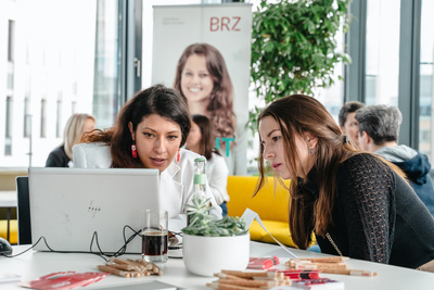 Zwei Frauen arbeiten konzentriert an einem Laptop, umgeben von Büromaterial in einem modernen Raum.