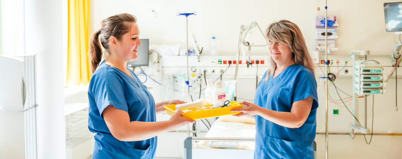 Zwei Pflegekräfte in blauen Uniformen übergeben sich ein Tablett mit medizinischen Utensilien in einem Krankenhauszimmer.