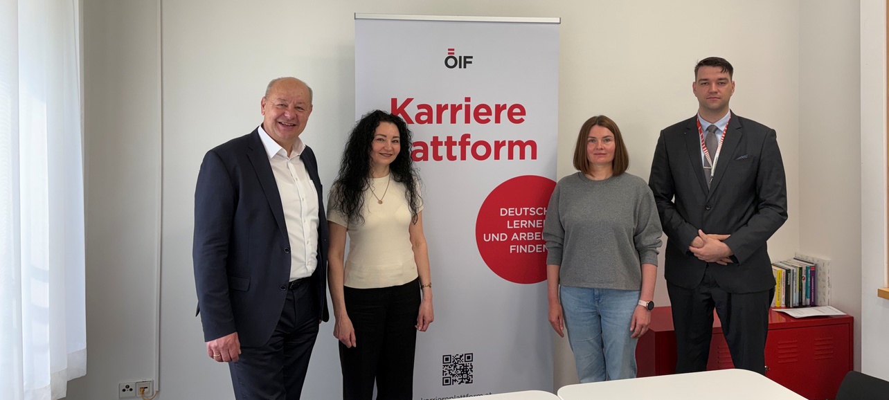 Vier Personen stehen vor einem Banner mit der Aufschrift "Karriereplattform. Deutsch lernen und Arbeit finden." im Innenraum. Die Personen bestehen aus einem älteren Mann, einer Frau mit lockigem Haar, einer Frau in einem grauen Pullover und einem Mann in einem Anzug.