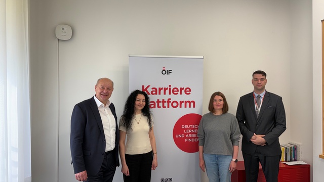 Vier Personen stehen vor einem Banner mit der Aufschrift "Karriereplattform. Deutsch lernen und Arbeit finden." im Innenraum. Die Personen bestehen aus einem älteren Mann, einer Frau mit lockigem Haar, einer Frau in einem grauen Pullover und einem Mann in einem Anzug.