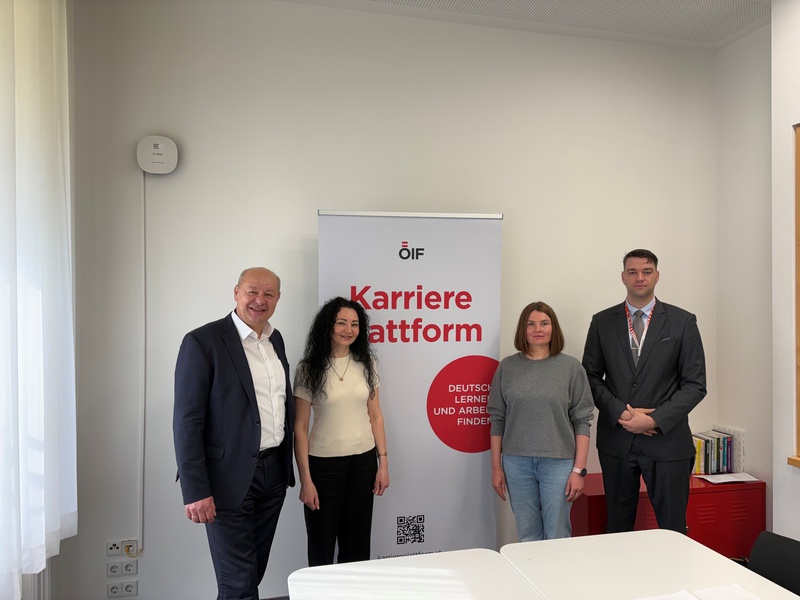 Vier Personen stehen vor einem Banner mit der Aufschrift "Karriereplattform. Deutsch lernen und Arbeit finden." im Innenraum. Die Personen bestehen aus einem älteren Mann, einer Frau mit lockigem Haar, einer Frau in einem grauen Pullover und einem Mann in einem Anzug.