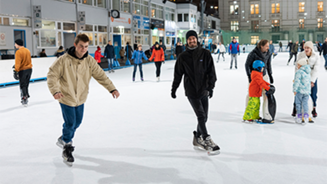 Zwei Männer laufen auf einem Eisbahn, umgeben von mehreren anderen Eisläufern und Kindern.