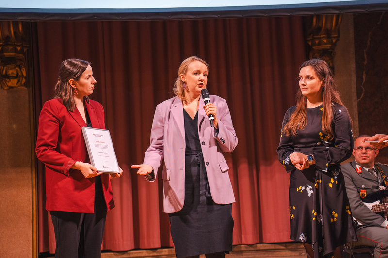 Drei Personen stehen auf einer Bühne; eine hält ein Zertifikat und spricht.