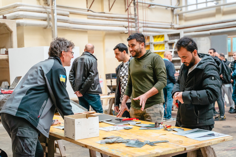 Gruppe von Menschen diskutiert an einem Tisch mit verschiedenen Materialien in einer Werkstatt.