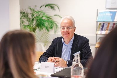 Ein älterer Mann mit Brille lächelt am Tisch während einer Besprechung.