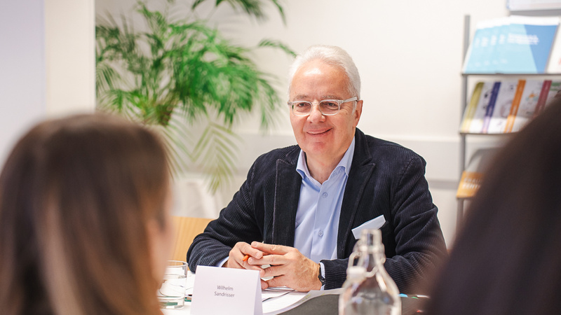 Ein älterer Mann mit Brille lächelt am Tisch während einer Besprechung.