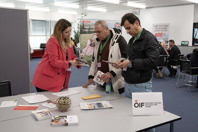 Eine Frau zeigt zwei Männern Informationsmaterial an einem Tisch in einem Veranstaltungsraum.