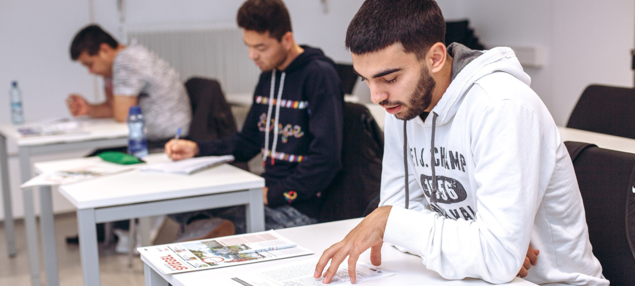 Drei junge Männer sitzen an Tischen und studieren Unterlagen in einem Klassenzimmer.