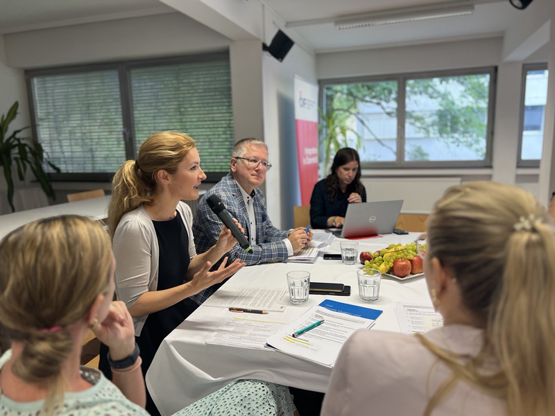 Eine Frau spricht während einer Besprechung, andere Teilnehmer hören aufmerksam zu und Notizen werden gemacht.