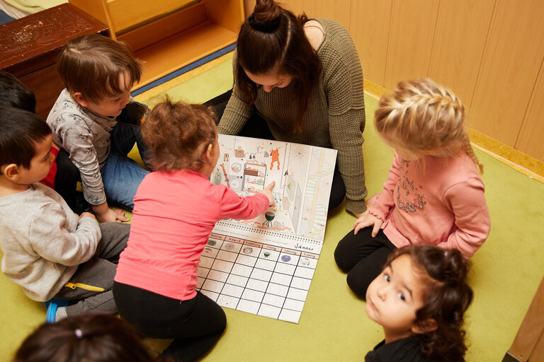 Eine Gruppe von Kindern und einer Erzieherin betrachtet gemeinsam ein buntes Arbeitsblatt auf dem Boden.