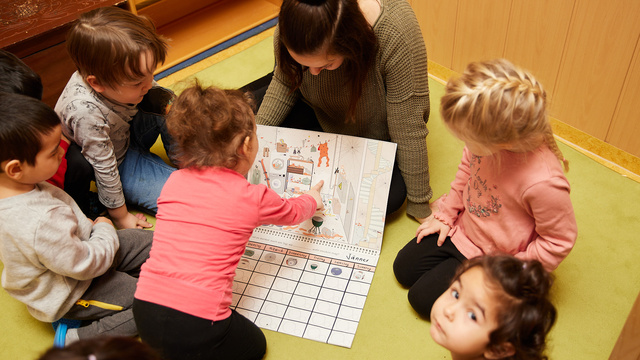 Eine Gruppe von Kindern und einer Erzieherin betrachtet gemeinsam ein buntes Arbeitsblatt auf dem Boden.
